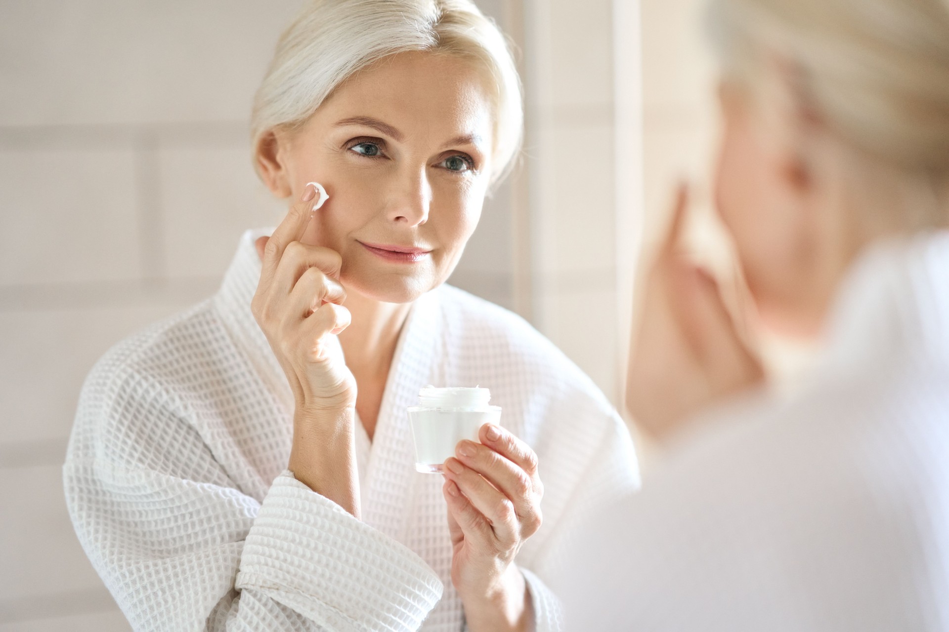 Happy smiling mid age woman looking at mirror applying cream. Skincare concept.